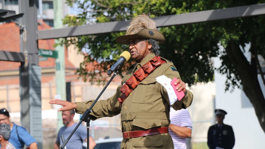 Ray Minniecon, dressed in military attire, stands on stage and speaks.