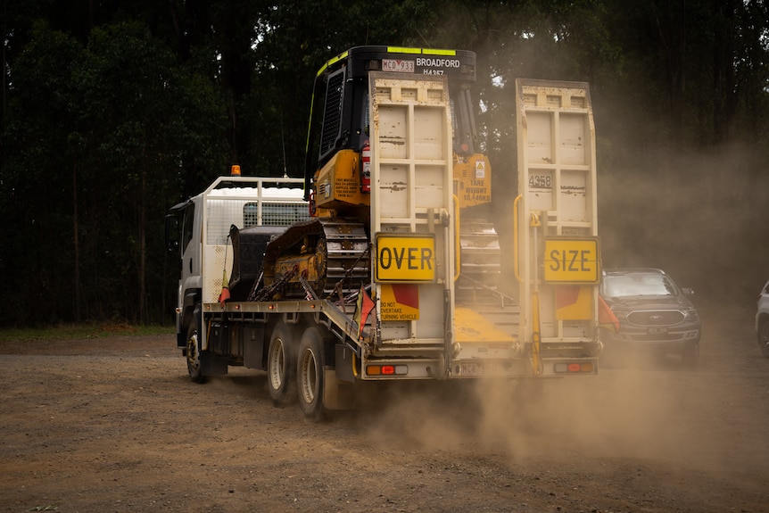 A truck carrying an excavator creating a dust cloud as it travels along a dirt road.