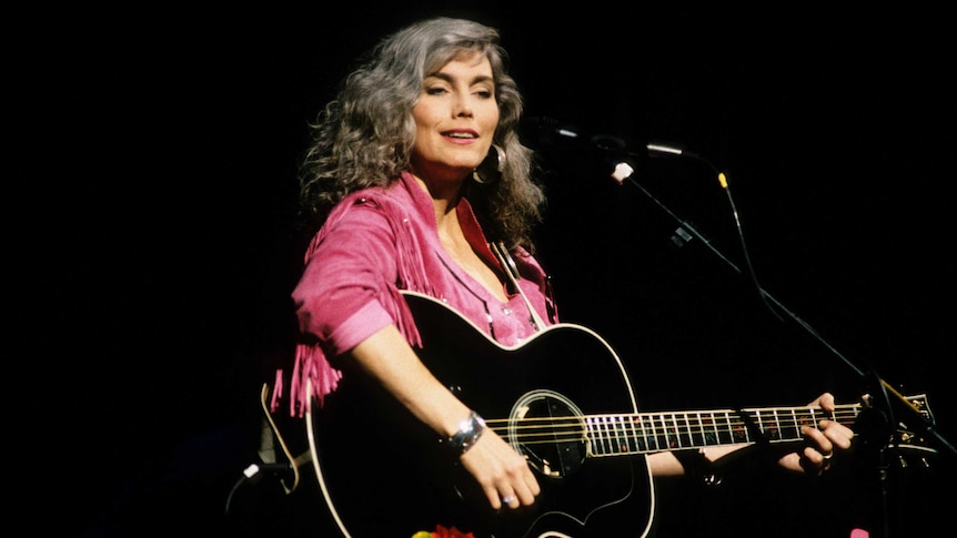 Emmylou is on stage wearing a pink fringed jacket and holding a black guitar. She's smiling.