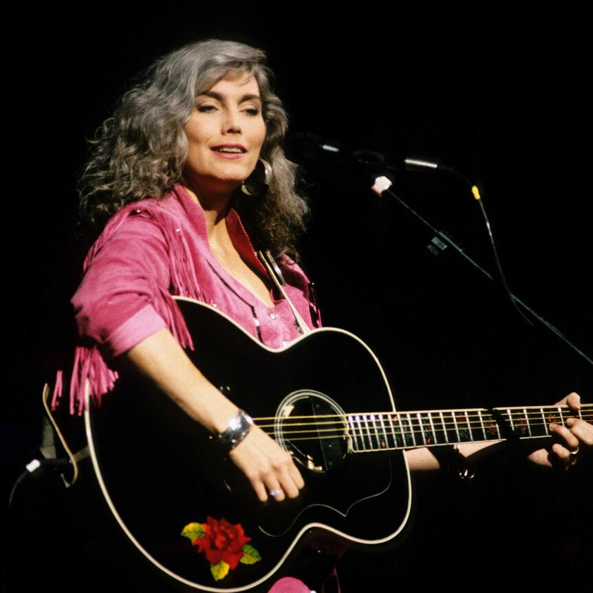 Emmylou is on stage wearing a pink fringed jacket and holding a black guitar. She's smiling.