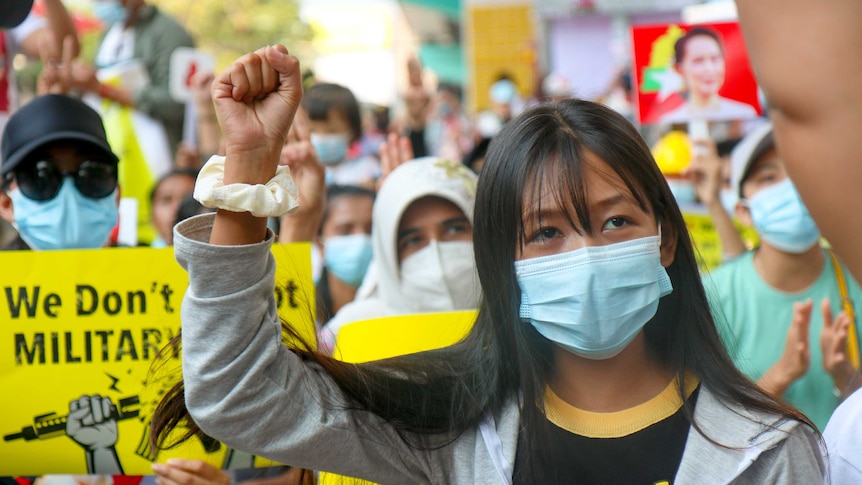 A woman with black hair and wearing a mask and jumper holds her fist in the air surrounded by a crowd of protesters.