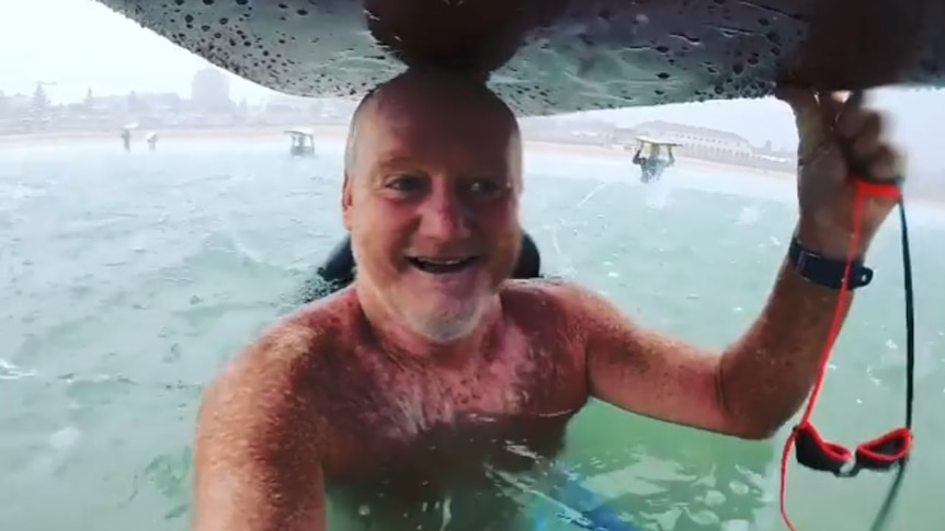 A man sheltering under a surfboard during a storm