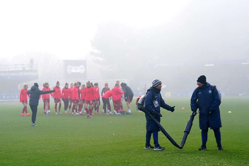 Groundskeepers with leaf blowers stand on the Kingsmeadow pitch as Liverpool players warm up for their match against Chelsea.