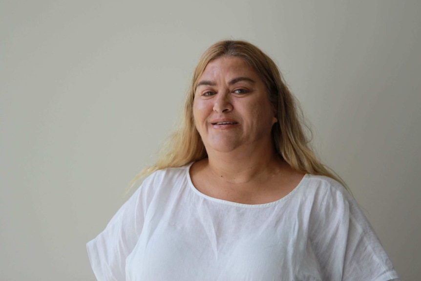 A woman with shoulder-length dyed blonde hair wears a white T-shirt and smiles 