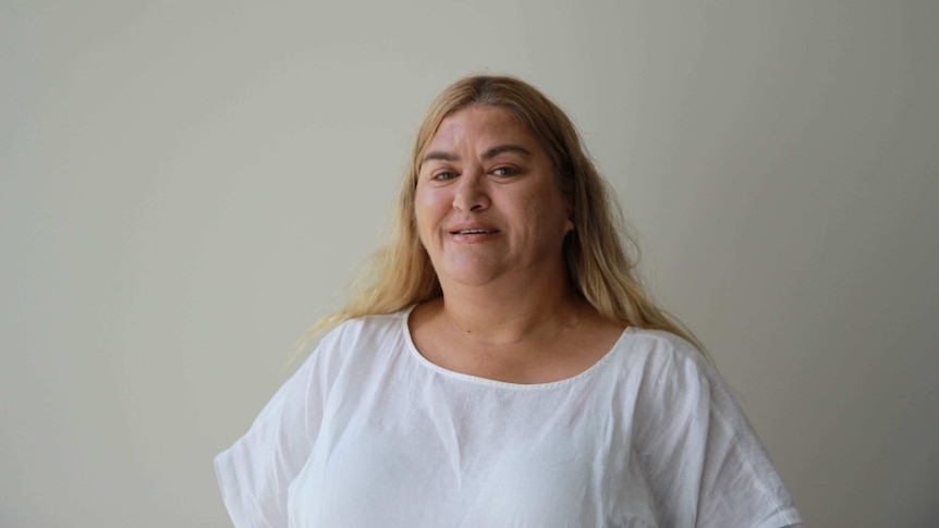 A woman with shoulder-length dyed blonde hair wears a white T-shirt and smiles 