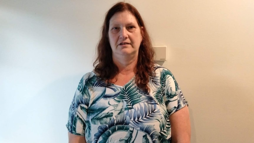 A woman in a blue and white top with dark brown hair stands in front of a white wall unsmiling.