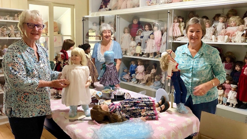 Ladies with grey hair and big smiles stand among hundreds of porcelain dolls.