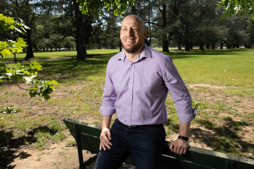 A man leans on a chair in a public park, smiling.