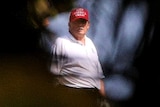 Donald Trump, wearing a white polo shirt and a red Keep America Great cap is framed by trees.