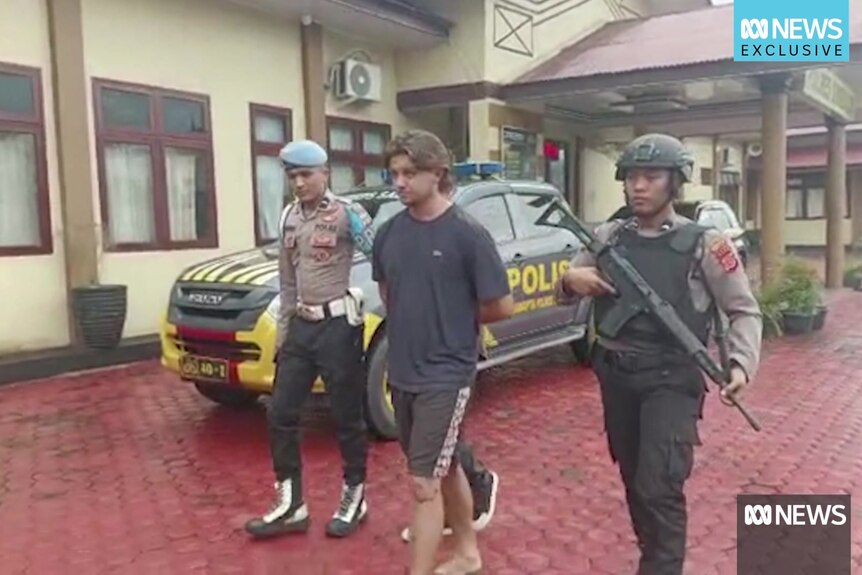 A young man in shorts in a t-shirt is escorted by two policeman in Indonesia.