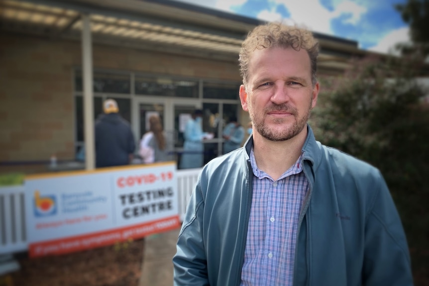 Mick Geary, wearing a blue jacket and shirt, stands in front of a suburban home on a sunny day.