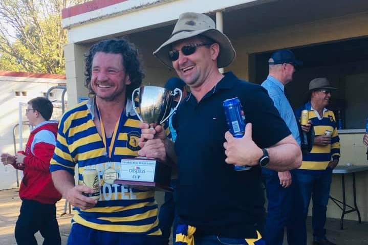 Two men holding beers and wearing rugby colours, each sporting big grins as they hold up a trophy.