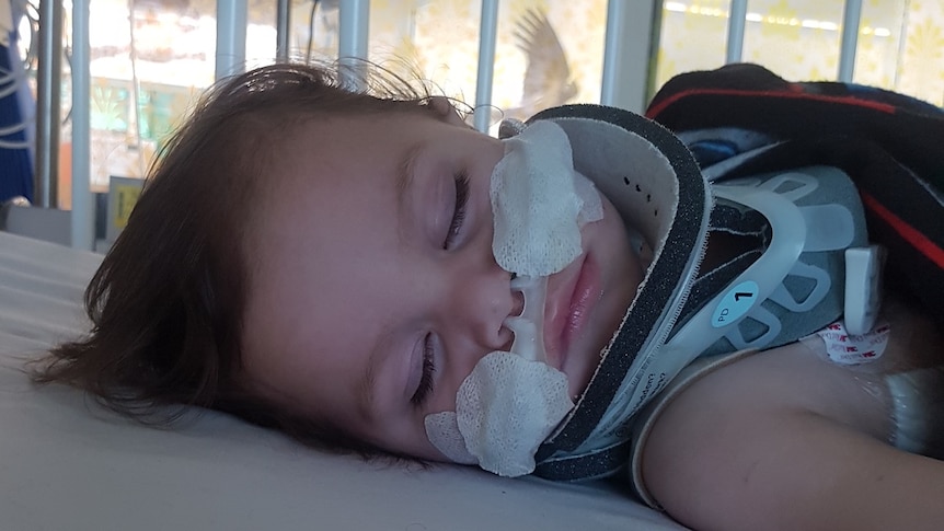 Toddler lies in hospital bed with neck brace.