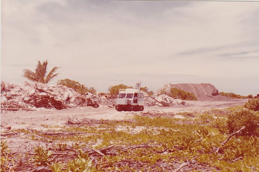 A white machine on treads drives along a dirt road past a pile of rubble.