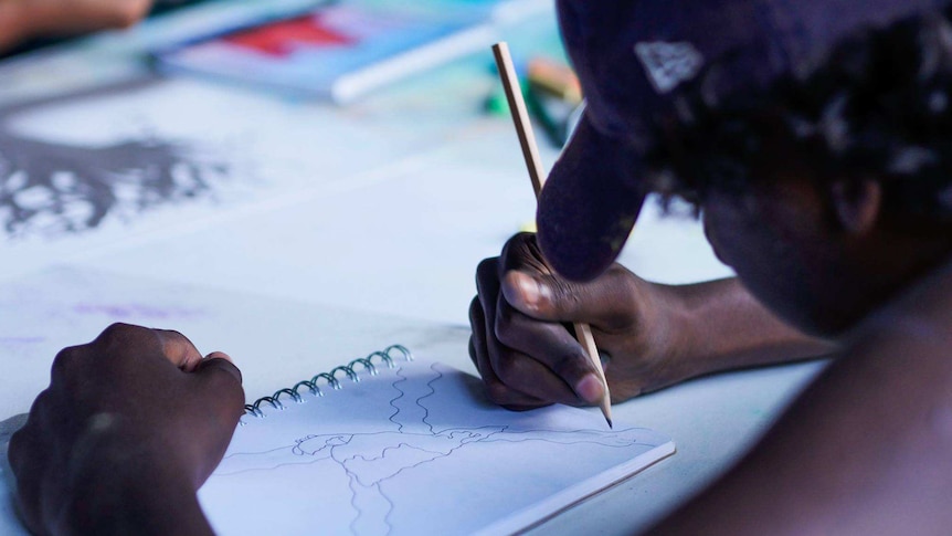 A student wearing a baseball cap is drawing with a pencil on a drawing notebook.