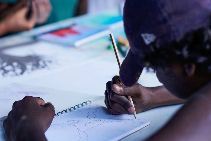 A student wearing a baseball cap is drawing with a pencil on a drawing notebook.