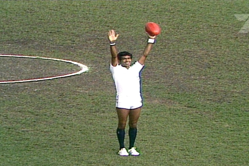 A man in a white shirt and white shorts stands in the middle of a football oval