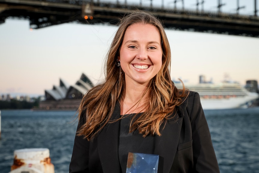 A young woman stands in front of sydney harbour holding a trophy