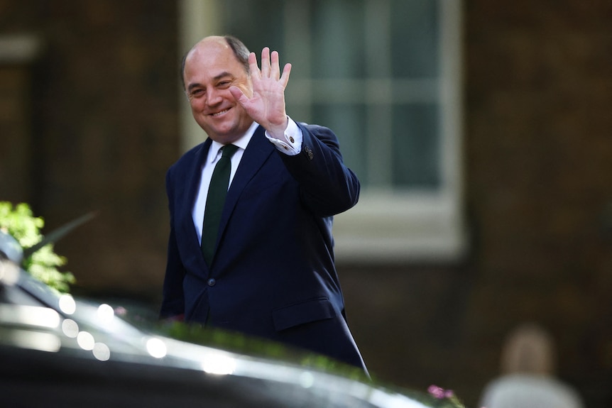 A man in a suit walks with a smile and casual wave