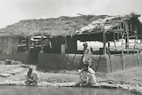 Three men in white robes sitting by an outback waterhole, next to a shack.