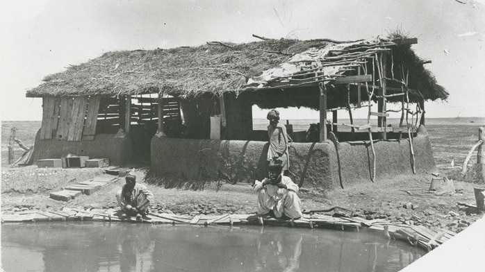 Three men in white robes sitting by an outback waterhole, next to a shack.
