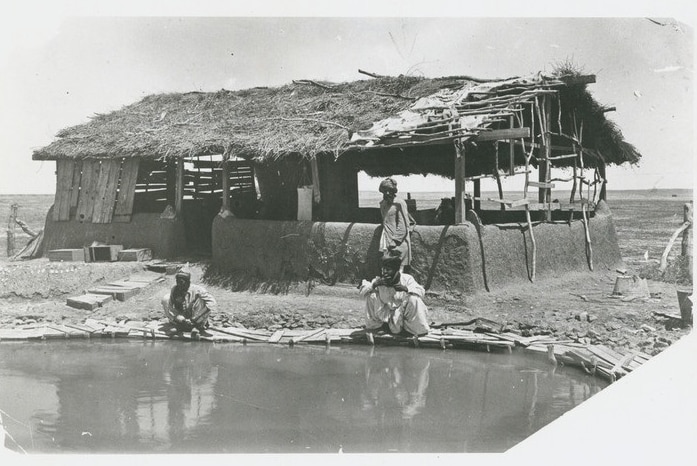 Three men in white robes sitting by an outback waterhole, next to a shack.