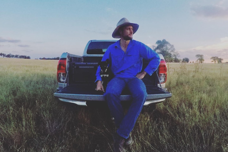 A man in an akubra sits in the back of an ute on a farm property.