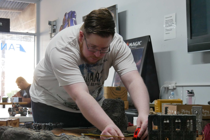 A man leans over a table covered with model buildings and figurines, measuring something with a tape measure.