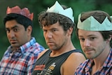 Three men sit unhappily next to each other wearing coloured paper crowns