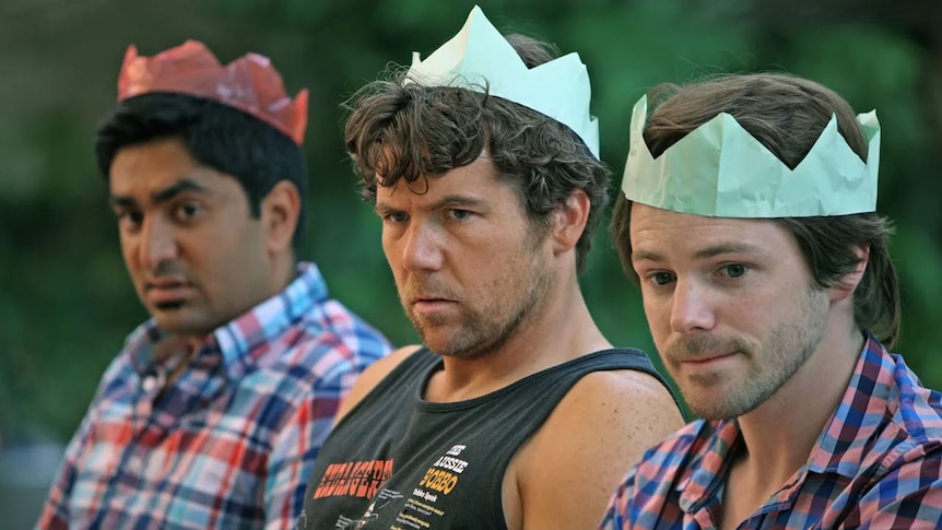 Three men sit unhappily next to each other wearing coloured paper crowns