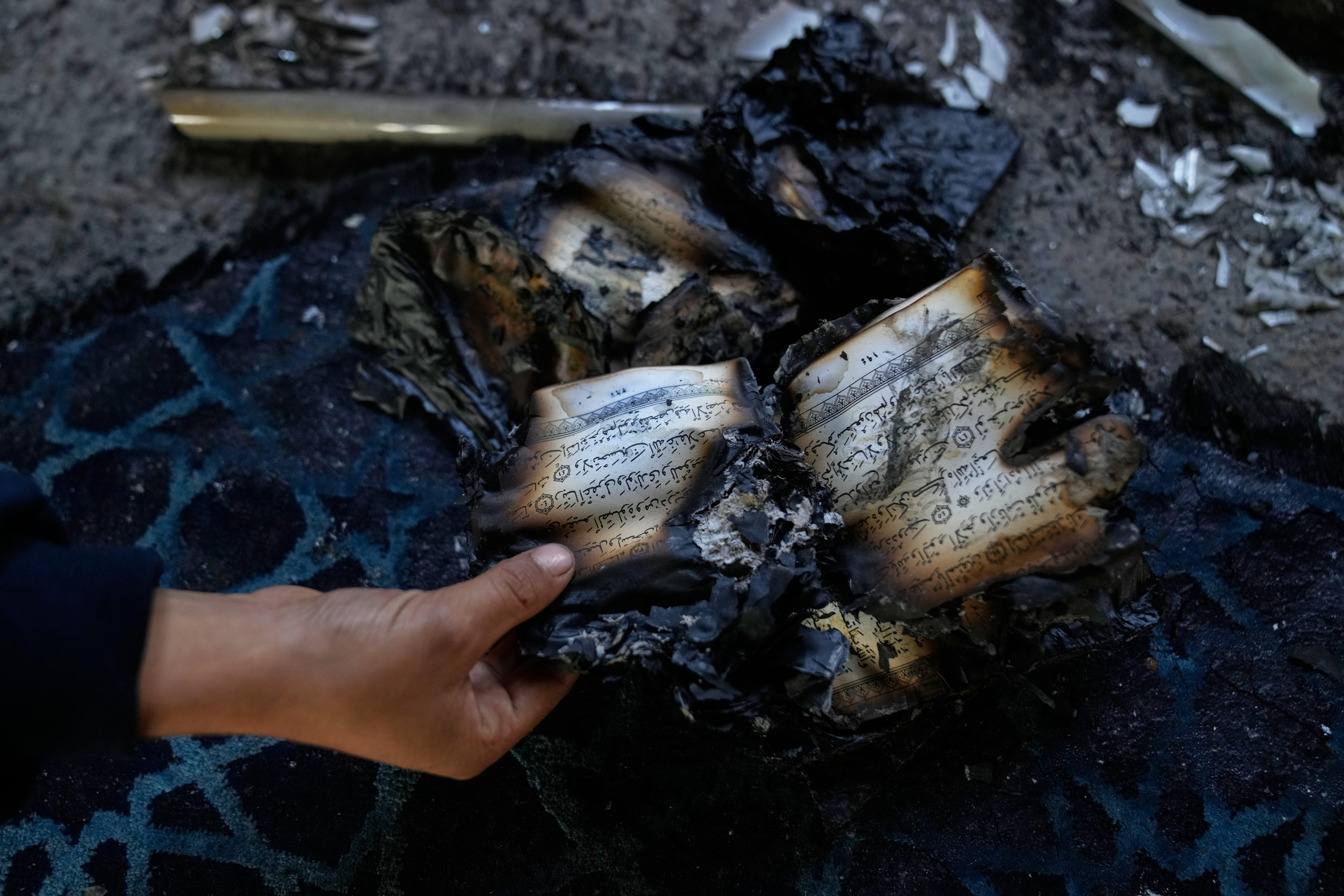 A charred Quran book with Arabic script held open by a hand, seen on the burnt pages.