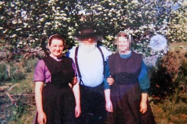 A young woman in an Amish dress and bonnet next to a man and woman, whose faces have been pixilated.