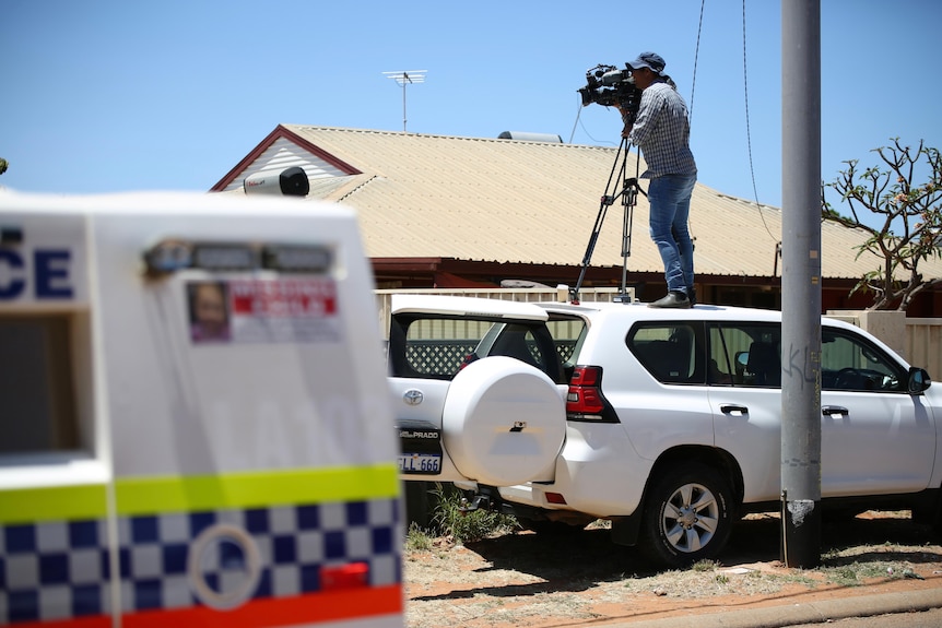 A camera man stands atop the roof of a four-wheel-drive filming, in the foreground is a police wagon.
