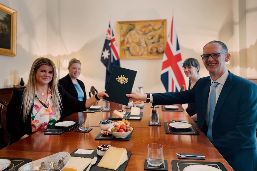 A woman and a man reach from opposites of a table to hold an official document, two other women sit on the wooden table behind.