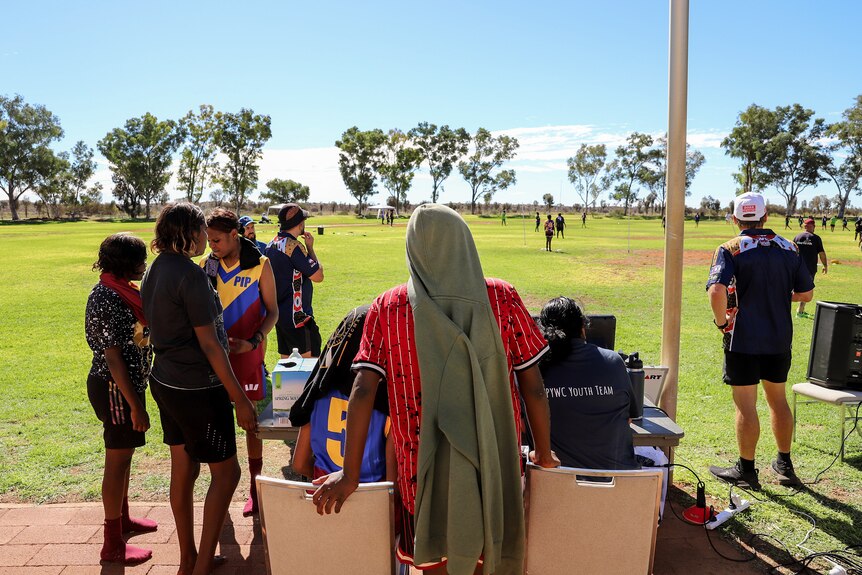A group of young Aboriginal women look out onto a green football ground where a small match is taking place