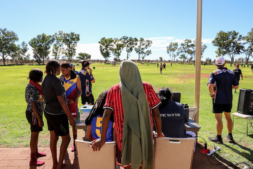A group of young Aboriginal women look out onto a green football ground where a small match is taking place