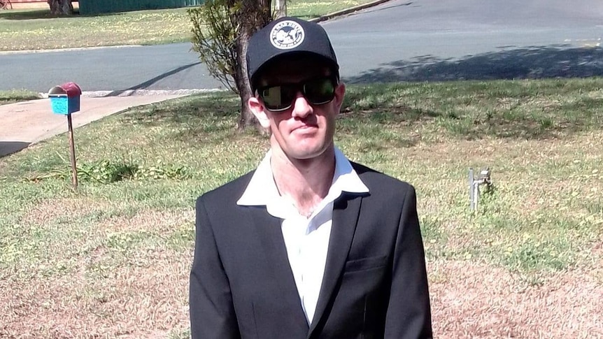 A caucasian man in a black suit jacket, white collared shirt, black cap and dark glasses stands on a front lawn.