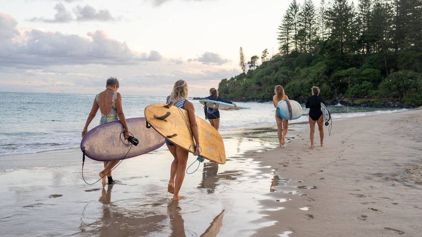 Five women with surfboards walk along a beach.