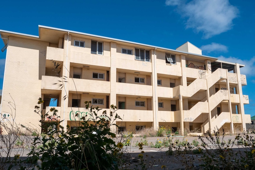 A cream-coloured apartment style building block stands in an empty carpark, covered with graffiti, windows smashed.