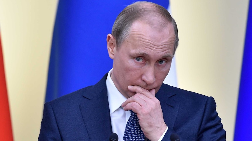 Vladimir Putin holds his hand to his face as he listens to someone asking a question.