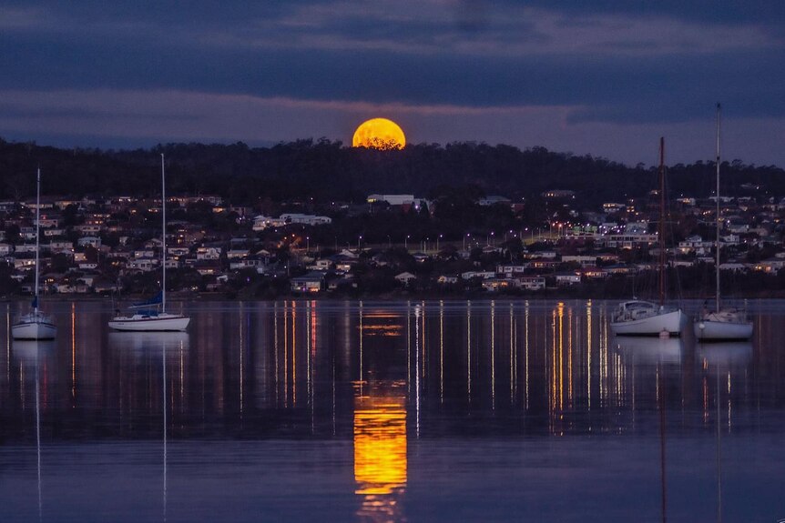 A full yellow moon rises behind mountains, casting a reflection on the water below.