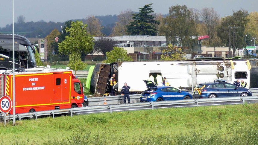 A bus lays on it's side next to a highway as emergency workers are seen on site.