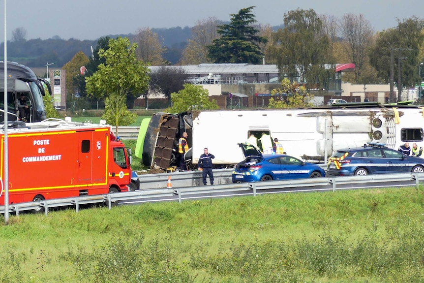 A bus lays on it's side next to a highway as emergency workers are seen on site.
