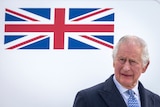 King Charles looks to the left of frame. Left of his head is the British flag.