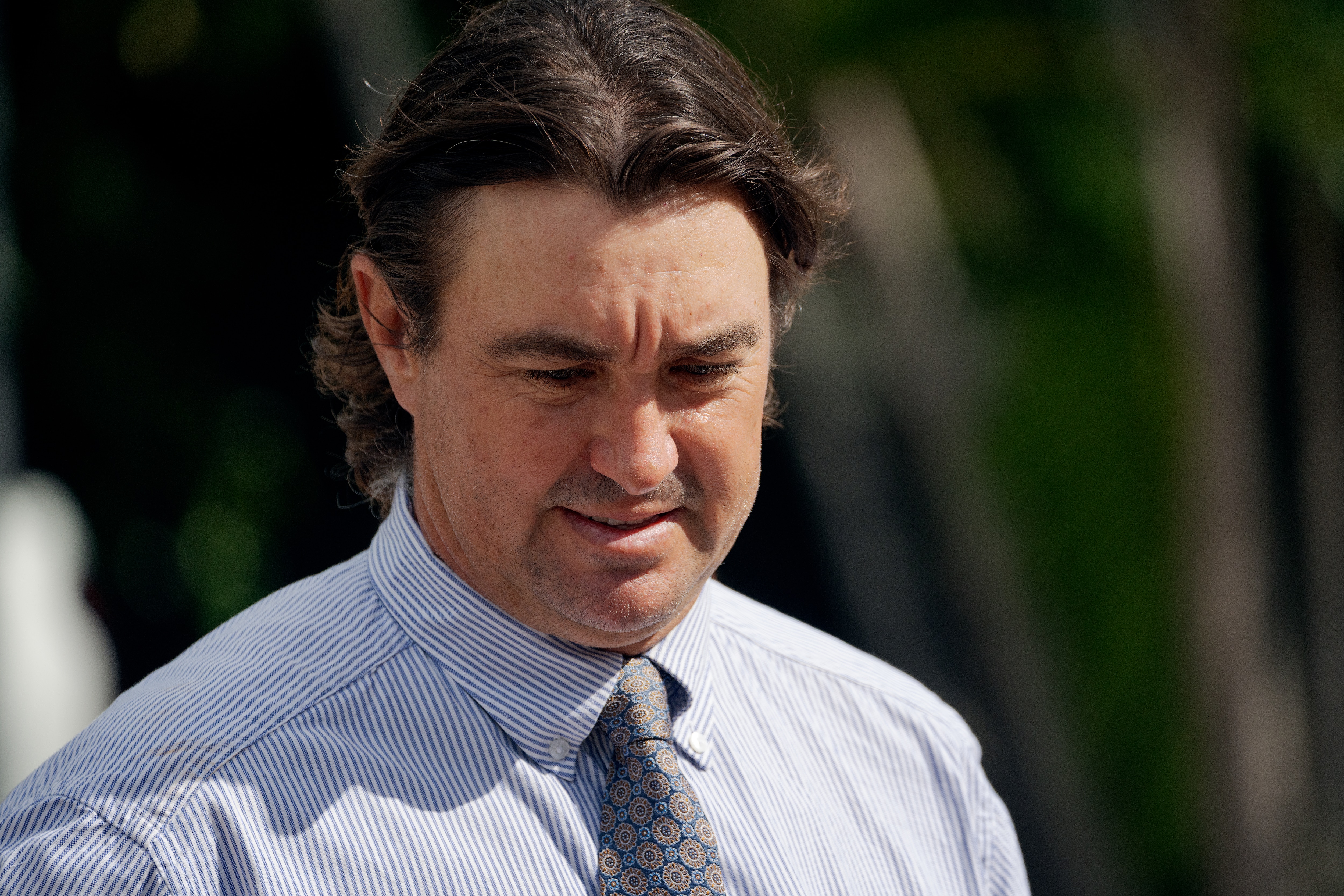 A close up of a white man, blue button up shirt, gray tie, brown hair at collar length, looking down.