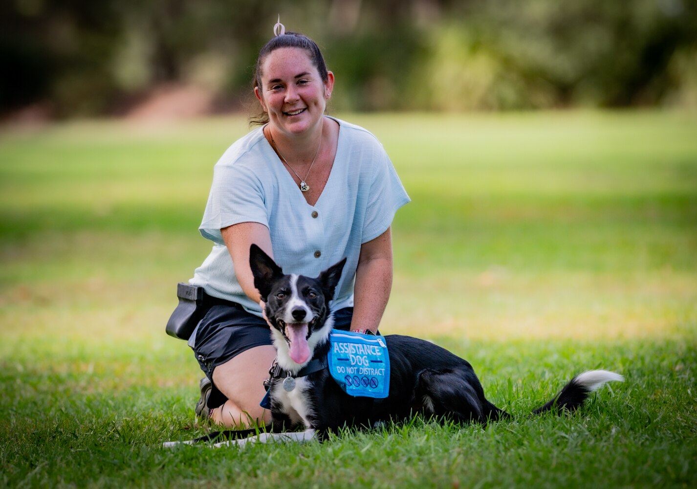 A young white woman sitting with her service dog in a park. The dog is a border collie and they are both smiling