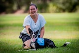 A young white woman sitting with her service dog in a park. The dog is a border collie and they are both smiling