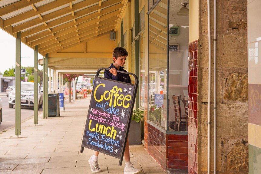 A boy in school uniform carries a cafe sign reading 'coffee and cake' inside a shop.