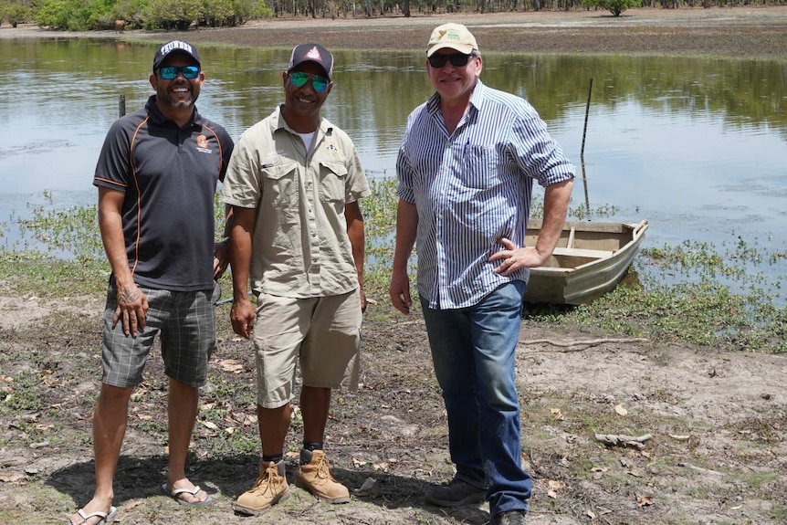 Three men wearing caps and sunglasses stand in front of a creek or river