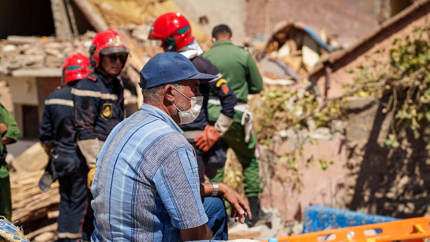 A man wearing a blue cap and striped shirt sits down and watches rescue workers clear rubble.